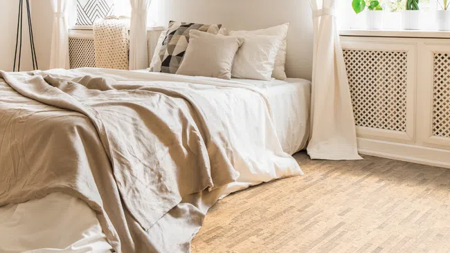 Bright bedroom with large windows, sheer curtains, neutral bedding, wooden floor, and potted plants on the windowsill.