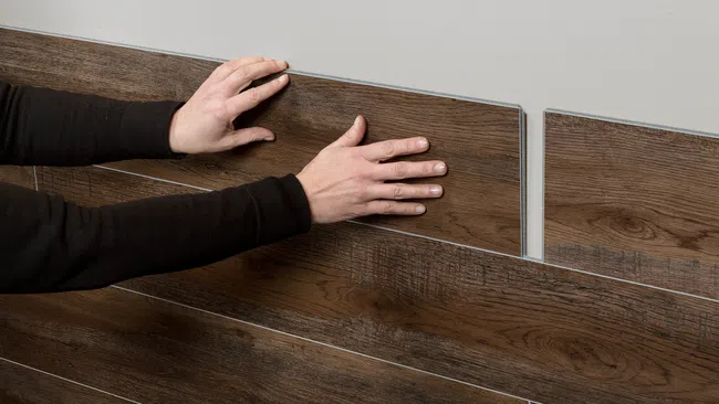 Hands installing dark wood laminate flooring panels on a light-colored wall or floor surface, aligning edges carefully.