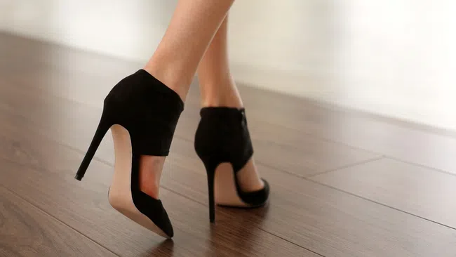 Close-up of feet wearing elegant black suede high heels walking on wooden floor indoors.