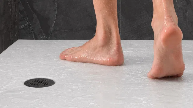 Wet bare feet standing on white textured shower floor near a circular drain with dark wall background.