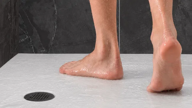 Wet bare feet standing on a white textured shower floor near a drain with black stone wall background.