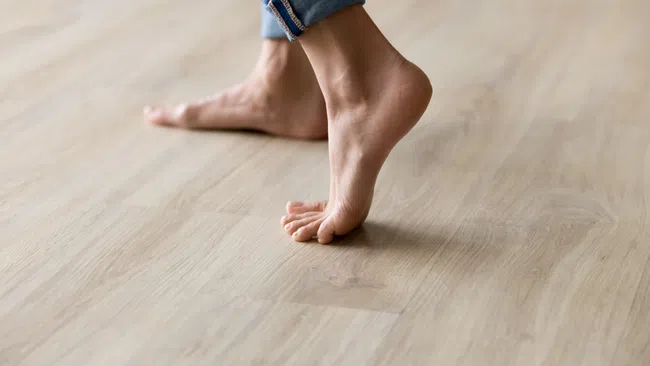 Close-up of bare feet walking on light wooden floor with rolled-up jeans visible on one leg.