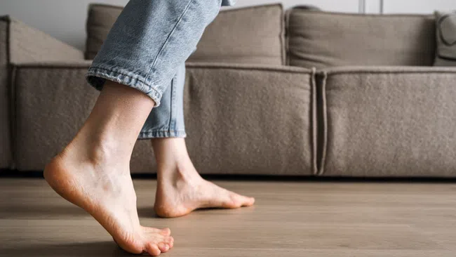 Person walking barefoot on wooden floor in living room with brown couch in background.