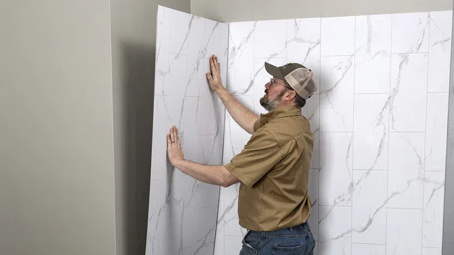 Man installing large white marble patterned wall panels in corner of room wearing casual clothes and cap.
