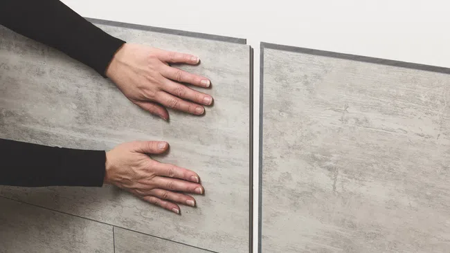 Hands installing large gray interlocking floor tiles with a concrete texture on a light floor.