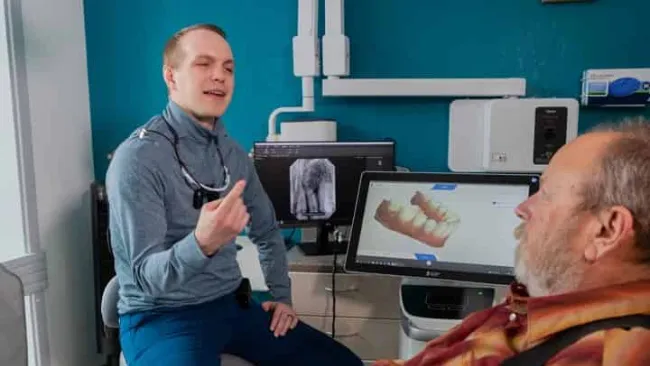 Dentist explaining dental scan results to a seated older male patient in a clinic with digital X-ray on screens.