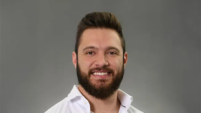 Smiling man with beard and styled hair wearing a white shirt against a gray background