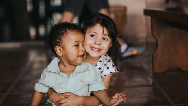 Two young children sitting on a tiled floor hugging and smiling in a cozy indoor setting