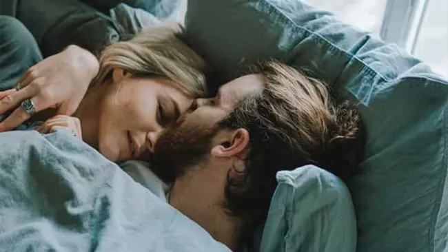 Couple cuddling peacefully in bed under blue blankets near a window with soft natural light.