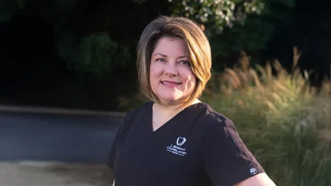 Smiling female healthcare professional in black scrubs outdoors with green foliage background.