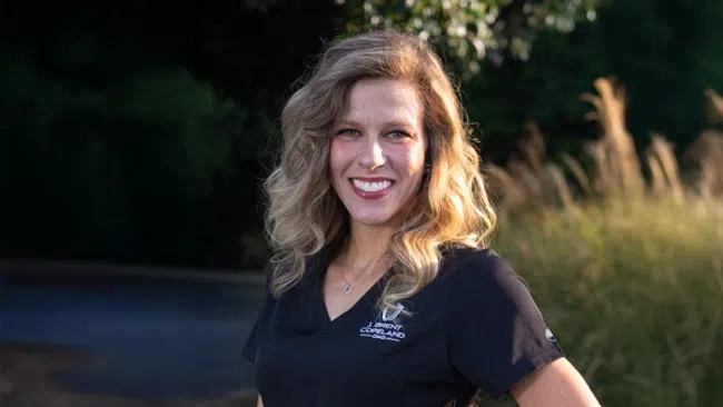 Smiling woman with blonde curly hair wearing a black scrubs top standing outdoors with greenery in the background