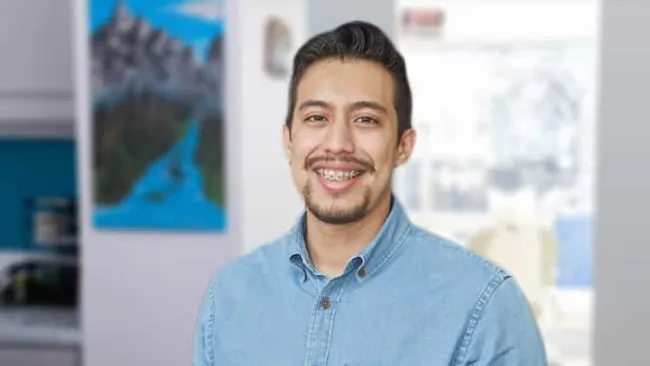 Smiling man with braces wearing a light blue denim shirt standing indoors with blurred background.