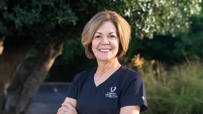 Smiling woman with short hair wearing a black J. Brent Copeland medical shirt outdoors with trees and greenery.