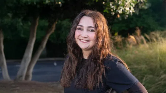 Young woman with long brown hair smiling outdoors in front of trees and grass during daytime