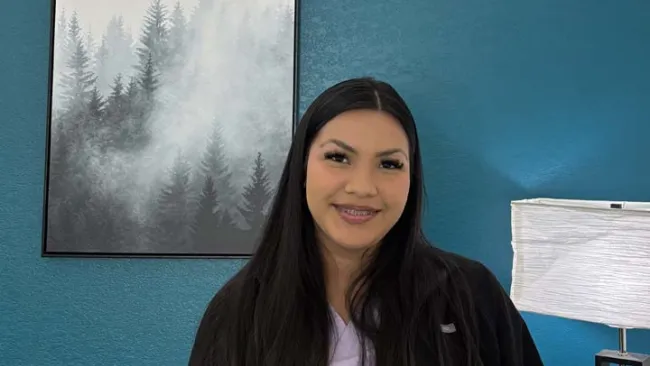 Smiling young woman with long dark hair and braces sits in blue room with forest painting and white lamp.