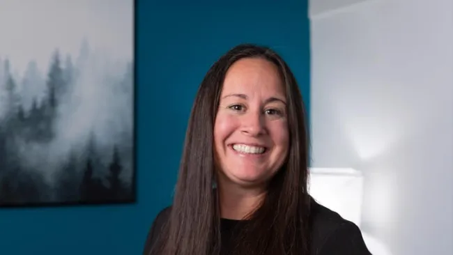 Smiling woman with long dark hair stands in a room with blue wall and misty forest wall art.