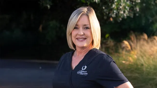 Smiling woman with blonde hair wearing a black dental uniform outdoors in natural light.