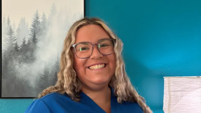 Smiling woman with wavy blonde hair and glasses wears blue scrubs standing against teal wall with lamp and forest painting.