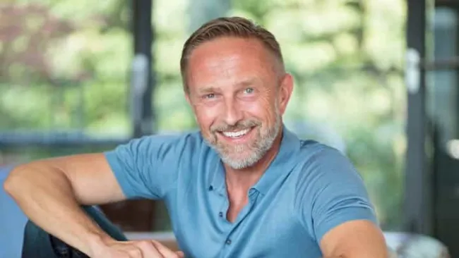 Smiling middle-aged man with beard wearing a blue polo shirt sitting comfortably indoors with natural light.