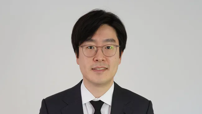 Man in formal attire with glasses, smiling against a plain white background.