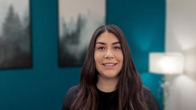 Smiling woman with long dark hair standing in front of teal wall with blurred forest art and lamp