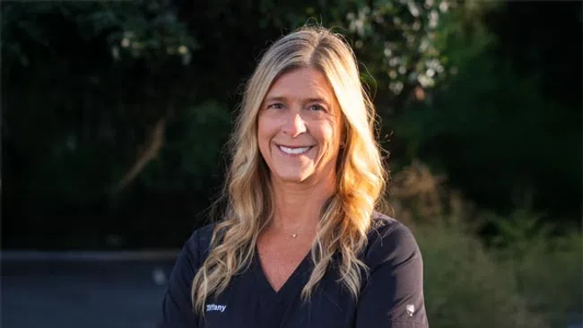 Smiling woman with long blonde hair in black medical scrubs standing outdoors with greenery in background