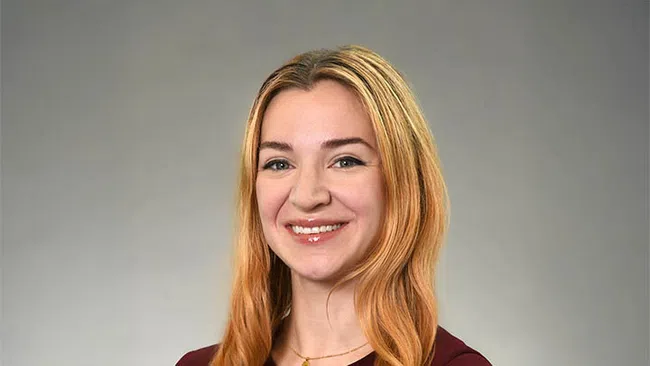 Smiling woman with long blonde hair wearing a maroon top against a neutral gray background