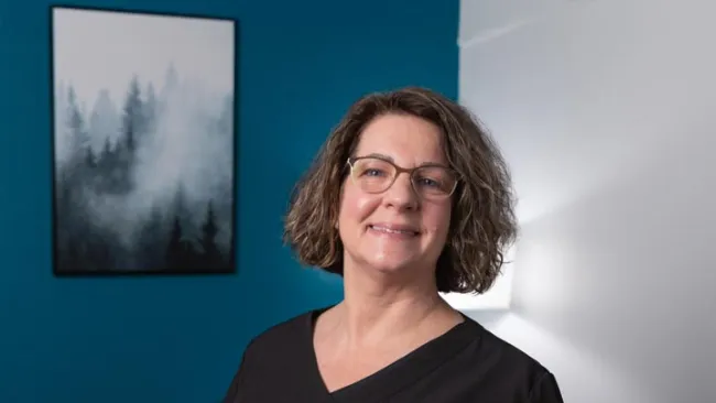 Smiling woman with glasses and curly hair wearing black shirt standing in a room with blue wall and forest painting.
