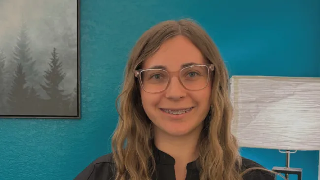 Smiling woman with glasses and braces in a black top against a teal wall with lamp and forest artwork.