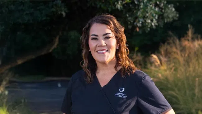 Smiling female dentist in black scrubs standing outdoors with greenery and sunlight in the background.