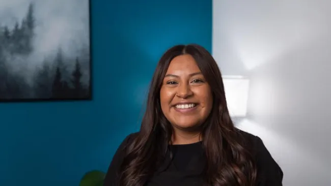 Smiling woman with long dark hair wearing black top against a blue and white background with wall art and lamp.