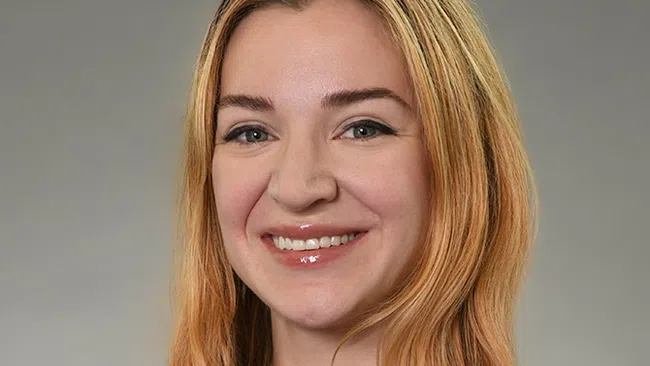 Smiling young woman with long blonde hair wearing a burgundy top and gold necklace on gray background.