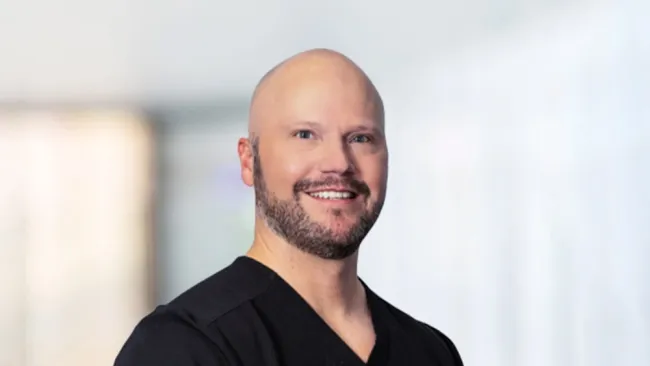 Smiling male healthcare professional in black scrubs with a blurred background.