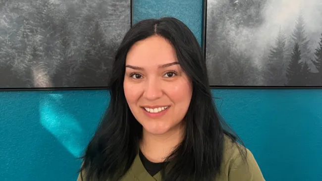 Smiling woman with long dark hair wearing olive green scrubs against a teal wall with forest paintings