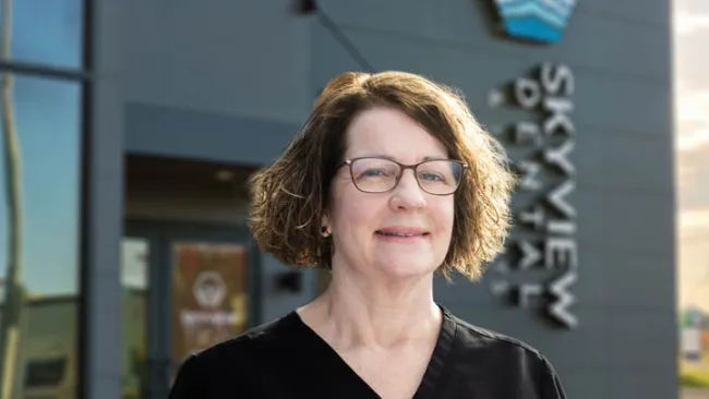 Middle-aged woman with glasses and curly hair standing outside Skyview Dental office wearing black scrubs.