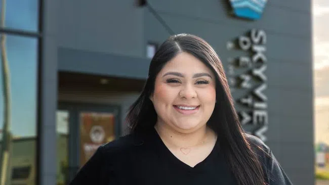 Smiling woman with long dark hair standing outside Skyview Dental clinic wearing a black shirt.