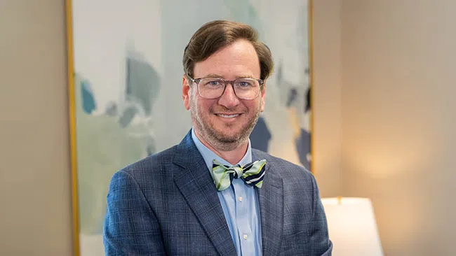 Man wearing glasses and a bow tie smiles in a professional indoor setting with abstract art and a lamp.