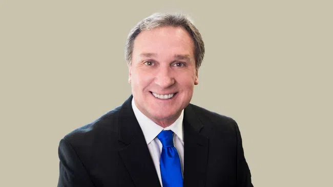 Smiling middle-aged man in black suit and blue tie against a plain beige background.
