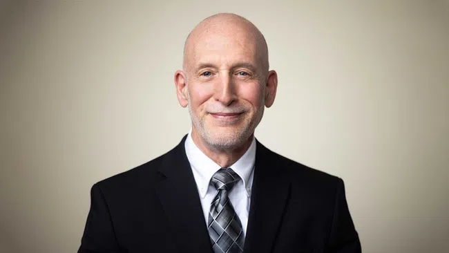 Smiling bald man wearing a black suit, white shirt, and gray patterned tie against a neutral background.