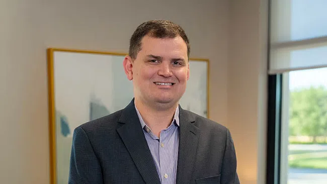 Smiling man in a dark blazer and striped shirt stands indoors near a window with blurred outdoor greenery.