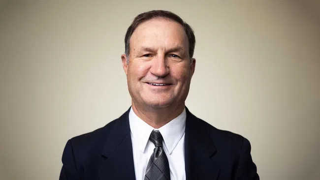 Middle-aged man smiling wearing a dark suit, white shirt, and patterned gray tie against a neutral background.