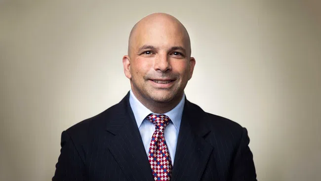 Smiling bald man in a dark pinstripe suit and patterned tie against a neutral background.