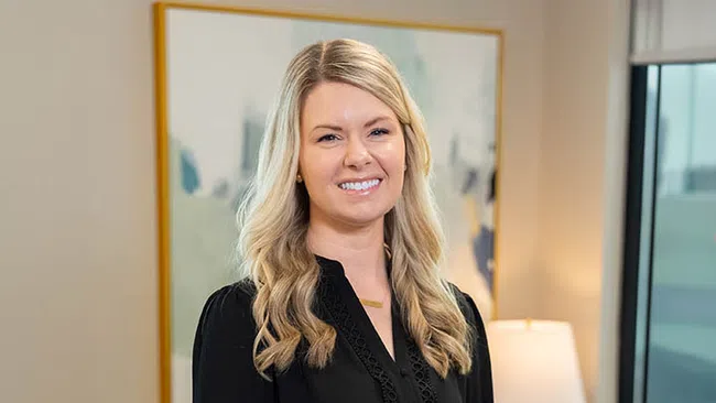 Smiling woman with long blonde hair wearing a black blouse standing in a modern office setting.
