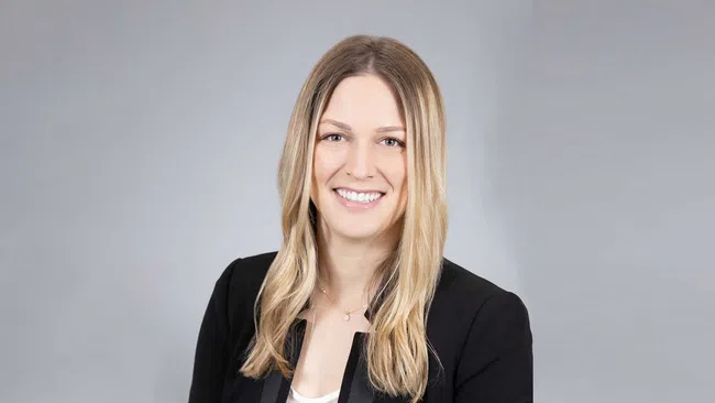 Smiling professional woman with blonde hair in black blazer posing against a light gray background.