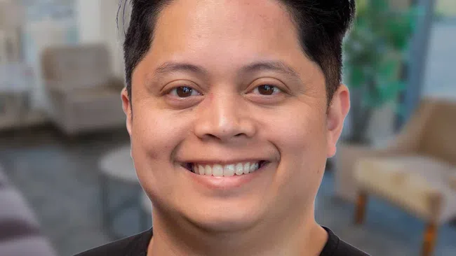 Smiling man with dark hair wearing a black V-neck shirt against a plain light background.