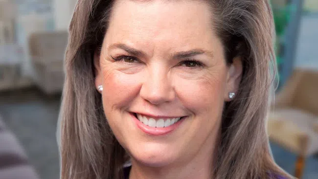 Smiling middle-aged woman with brown hair wearing a blue top and a pendant necklace against a neutral background