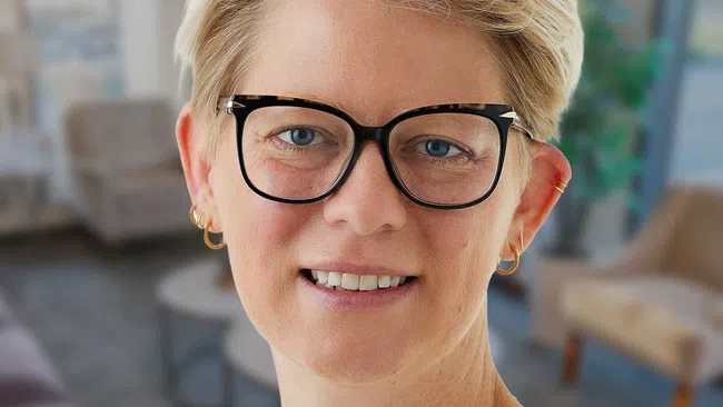 Smiling woman with short blonde hair wearing black glasses and a white lace detailed top against a light background.