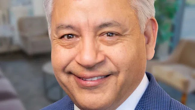 Smiling middle-aged man with gray hair wearing a blue suit and pink patterned tie on a plain background