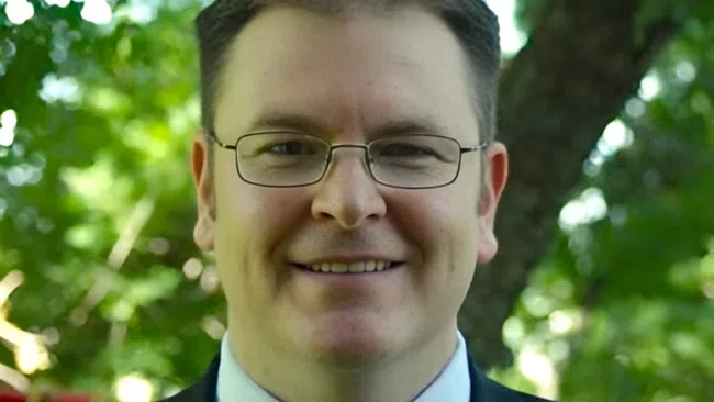 Smiling man in glasses wearing a suit with a yellow tie outdoors with green leafy background