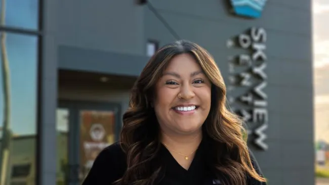 Smiling woman with wavy hair standing outside Skyview Dental clinic in black uniform.